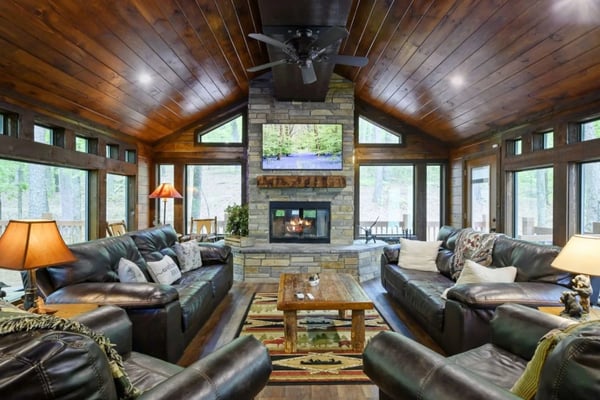 Spacious wood-paneled living room with stone fireplace, black leather seating, and forest views through large windows
